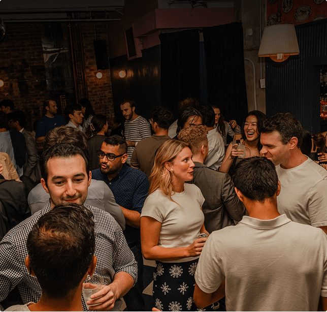 A crowded indoor party with people socializing, holding drinks, and engaging in conversation in a dimly lit room.